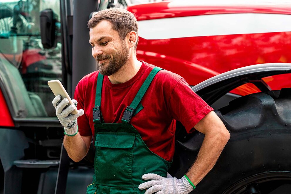 Ein männlicher Landwirt steht neben einem Traktor, telefoniert mit seinem Handy und lächelt. 