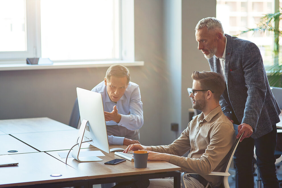 Drei männliche Mitarbeiter, die auf einen PC-Bildschirm schauen und ein Projekt besprechen, Geschäftsleute, die im Coworking Space zusammenarbeiten