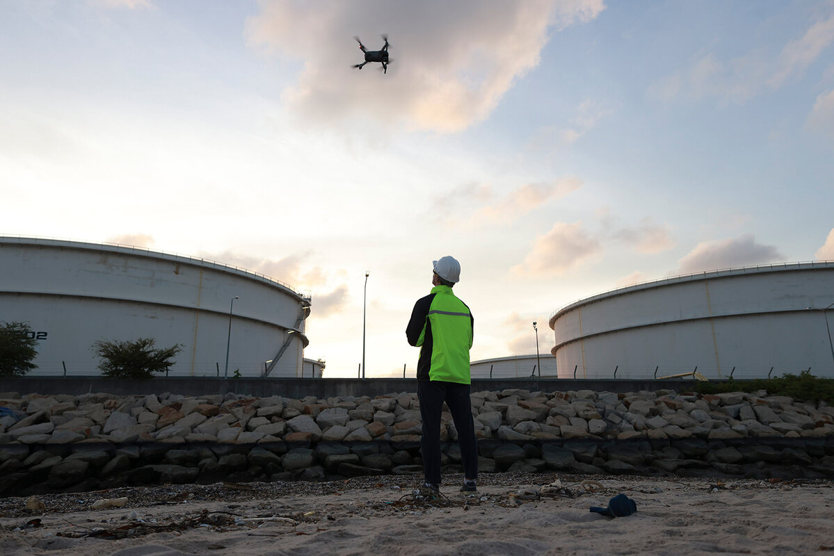 Leitender Energieingenieur steuert eine Drohne über einem Öltanklager. Videoüberwachung oder industrielle Inspektion in der Raffinerie. 