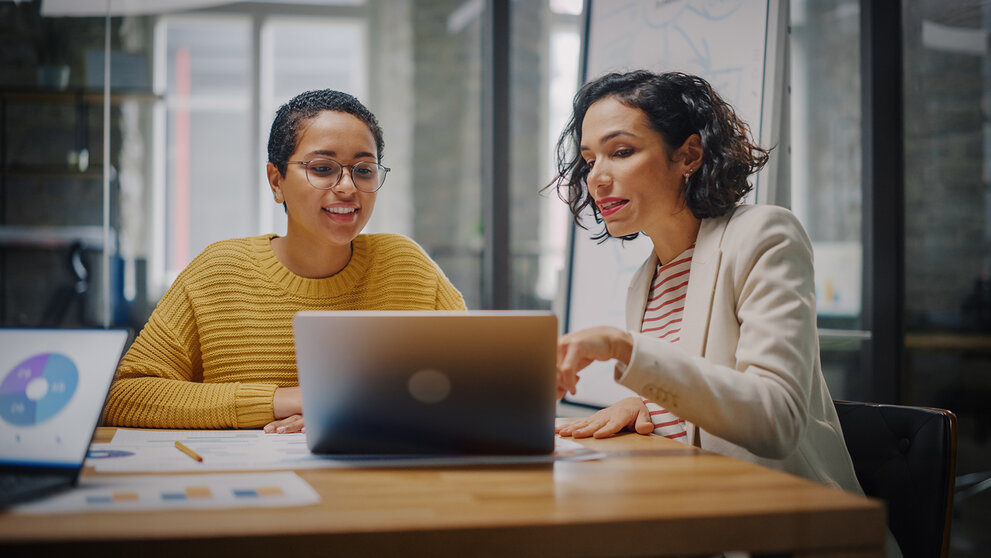 Zwei Frauen unterhalten sich in einem Besprechungsraum mit Glaswänden in einer Agentur. Eine Kreativdirektorin und eine Projektmanagerin vergleichen in einem Büro die Geschäftsergebnisse auf einem Laptop sowie App-Entwürfe.