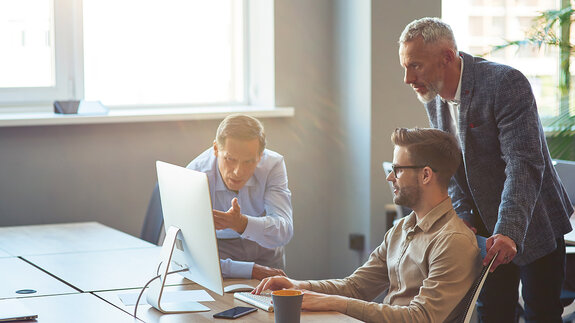 Drei männliche Mitarbeiter, die auf einen PC-Bildschirm schauen und ein Projekt besprechen, Geschäftsleute, die im Coworking Space zusammenarbeiten
