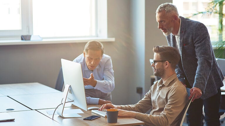 Drei männliche Mitarbeiter, die auf einen PC-Bildschirm schauen und ein Projekt besprechen, Geschäftsleute, die im Coworking Space zusammenarbeiten