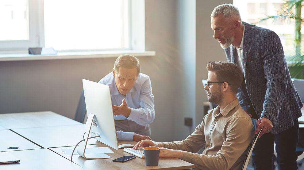Drei männliche Mitarbeiter, die auf einen PC-Bildschirm schauen und ein Projekt besprechen, Geschäftsleute, die im Coworking Space zusammenarbeiten