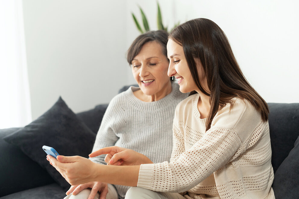 Eine erwachsene Tochter hilft ihrer Mutter, herauszufinden, wie man ein Smartphone benutzt, zwei Mehrgenerationen-Frauen sitzen auf der Couch und schauen auf den Handy-Bildschirm