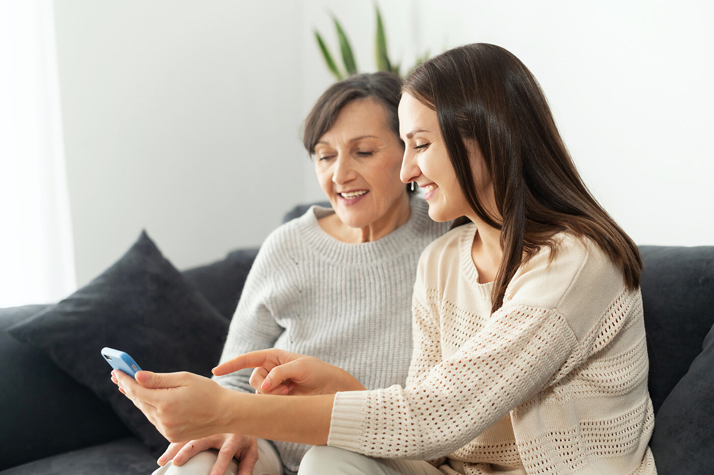 Eine erwachsene Tochter hilft ihrer Mutter, herauszufinden, wie man ein Smartphone benutzt, zwei Mehrgenerationen-Frauen sitzen auf der Couch und schauen auf den Handy-Bildschirm