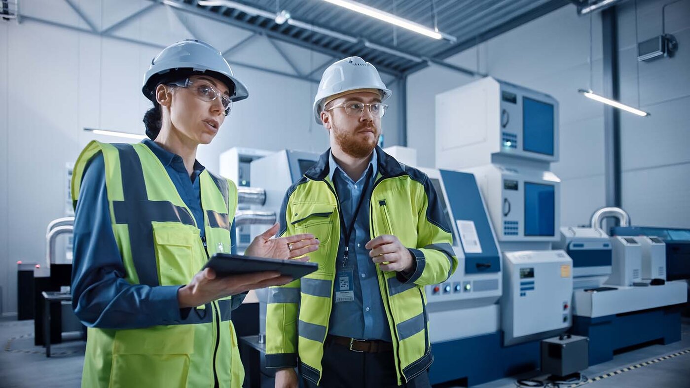 Moderne Fabrik: Projektmanagerin und Ingenieur stehen in Schutzwesten und Schutzhelmen in der Werkstatt, unterhalten sich, bedienen ein Tablet und überwachen die CNC-Fertigungsstraße