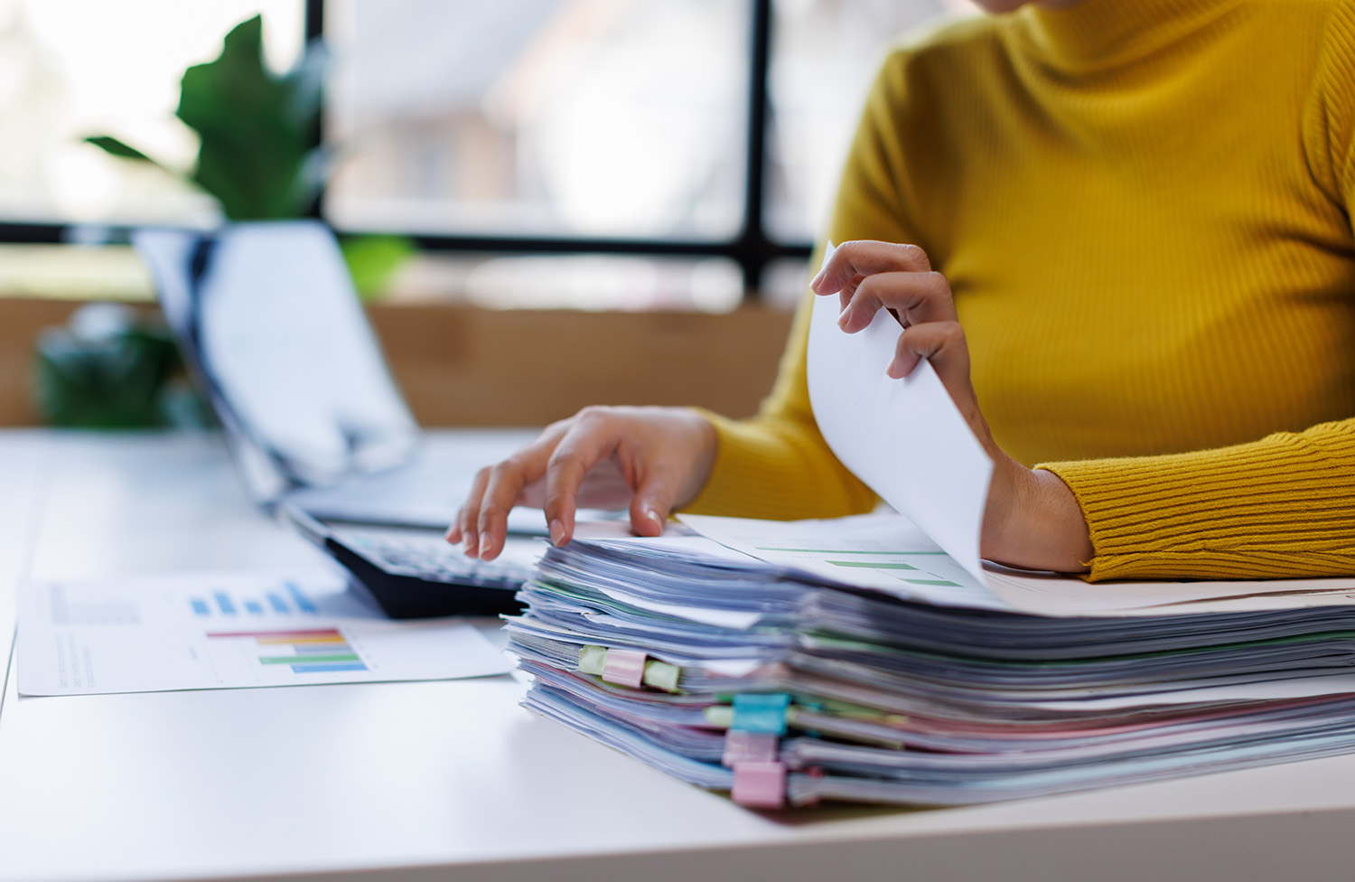 Eine Geschäftsfrau arbeitet an einem überfüllten Schreibtisch im Büro und durchsucht Stapel von Papierakten, um unvollständige Dokumente in Ordnern zu finden