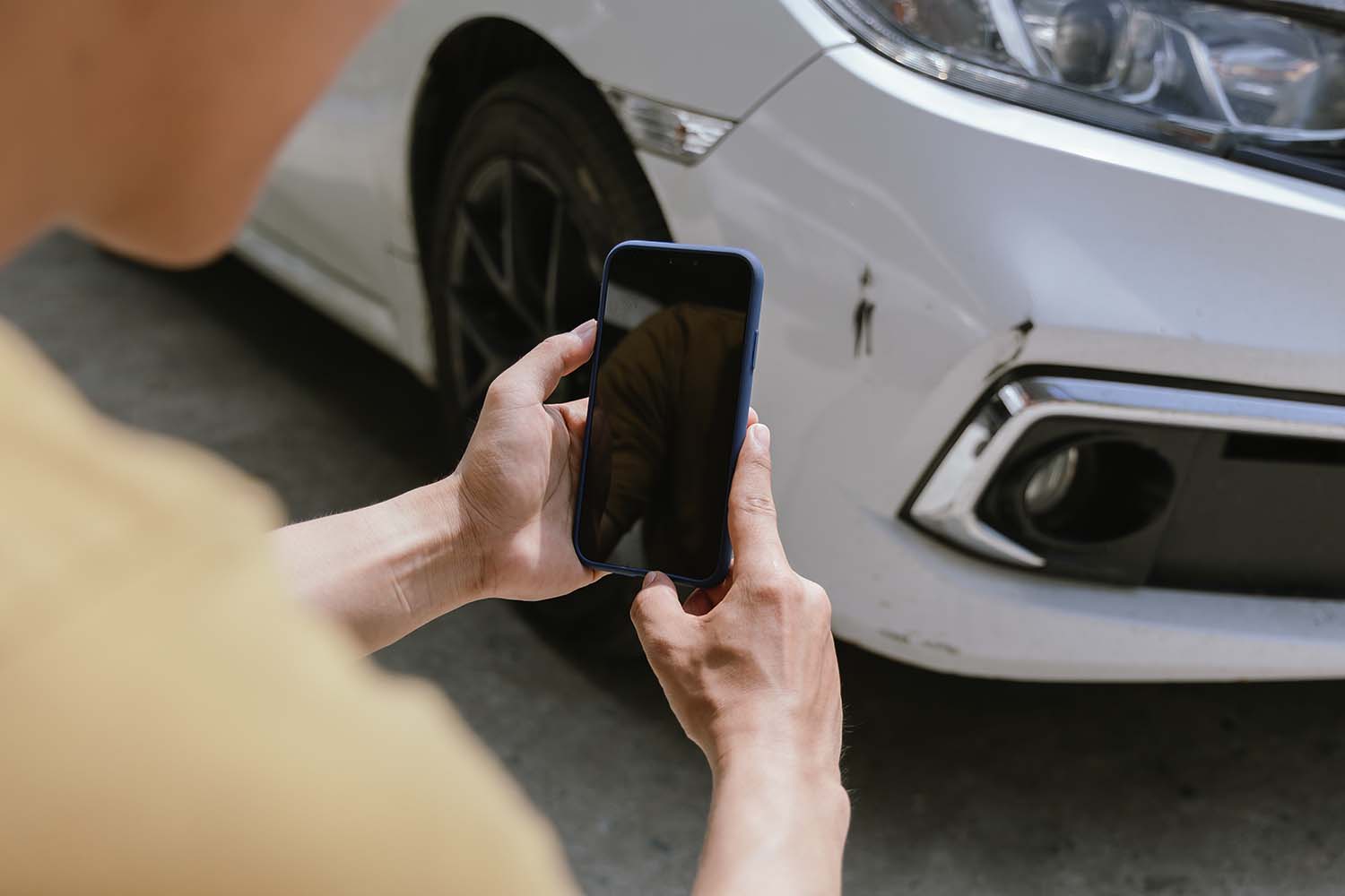 Ein junger Mann hält ein Smartphone in der Hand und fotografiert sein nach einem Unfall verbeultes Auto. Ein Versicherungsagent mit einem Klemmbrett begutachtet den Schaden und erstellt einen Schadensbericht.
